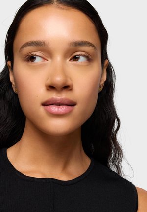 Młoda kobieta z długimi ciemnymi włosami i gładką cerą, nosząca małe kolczyki i czarny top bez rękawów, patrząca w lewo.