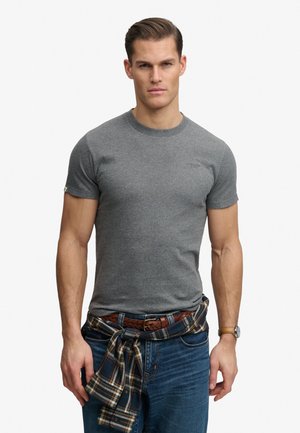 Mężczyzna w dopasowanym szarym krótkim rękawem, niebieskich dżinsach, z koszulą w kratę zawiązaną wokół talii oraz brązowym zegarkiem na lewym nadgarstku.