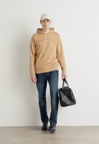 Felpa con cappuccio marrone con la scritta "GANT 1949" stampata in viola, abbinata a jeans blu e scarpe nere, mentre tiene una borsa nera. Sfondo bianco.