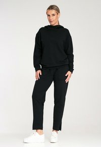 Czarny rozpinany hoodie o luźnym kroju, z wysokim kołnierzem i długimi rękawami, w połączeniu z czarnymi spodniami z kieszenią i bocznymi rozporkami. Białe sneakersy.