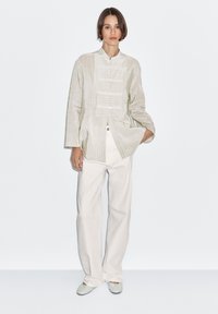 Jasna koszula zapinana na guziki z pionowymi paskami, z wysokim kołnierzem i długimi rękawami. Noszona z białymi spodniami i jasnymi butami.