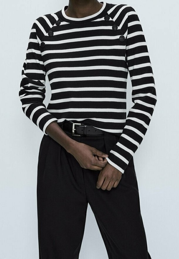 Personne portant une chemise à manches longues rayée horizontalement noir et blanc avec des boutons près des épaules, associée à un pantalon noir taille haute et une ceinture.