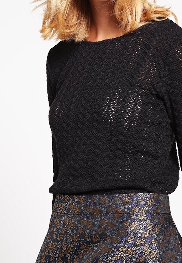 Personne blonde portant un haut noir texturé à manches longues associé à une jupe brillante à motif floral dans des couleurs sombres et métalliques.
