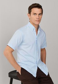 Homme assis sur un tabouret noir portant une chemise bleu clair à manches courtes boutonnée et un pantalon marron foncé sur un fond neutre.