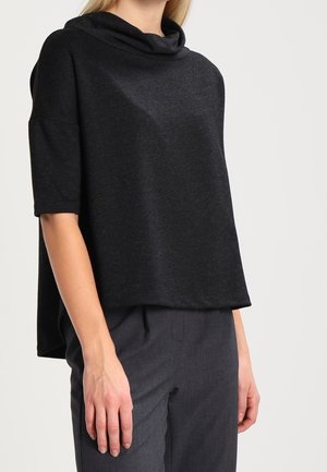 Kobieta w luźnej czarnej bluzce z krótkim rękawem i kołnierzem typu komin oraz ciemnoszarych spodniach, pokazana od szyi do bioder na jednolitym tle.