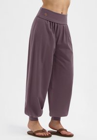 Eine Person trägt lockere, hochgeschnittene lilafarbene Hosen mit gerafften Bündchen und braunen Flip-Flops vor einem weißen Hintergrund.