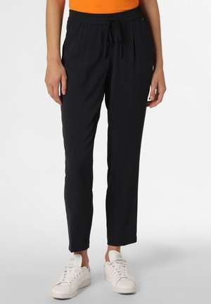 Kobieta w czarnych spodniach z sznurkiem i białych tenisówkach stoi na tle jednolitego tła.