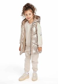 Gilet matelassé argent métallisé avec capuche, sweat-shirt beige et sweat-pants assortis. Baskets blanches avec accents beiges et semelles marron clair.