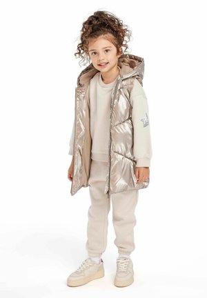 Gilet metallizzato argento imbottito con cappuccio, felpa beige e pantaloni della tuta coordinati. Sneakers bianche con dettagli beige e suole marrone chiaro.