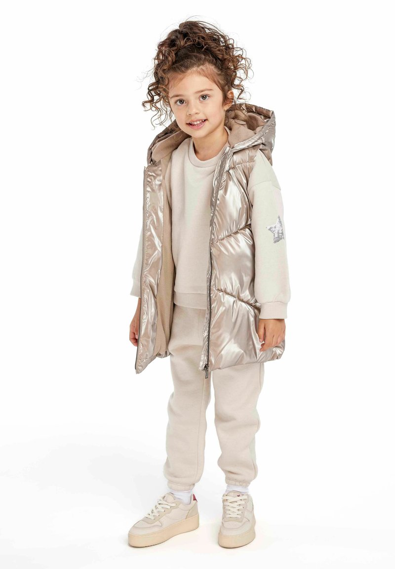Gilet matelassé argent métallisé avec capuche, sweat-shirt beige et sweat-pants assortis. Baskets blanches avec accents beiges et semelles marron clair.