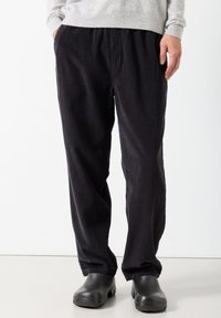 Osoba koja nosi široke tamne pamučne hlače s rukama u džepovima i crne obuće na svijetlom podu.