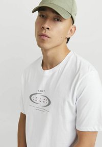 Hvid bomulds t-shirt med korte ærmer, der har et grå og burgunder logo print i midten. Modellen bærer en lysegrøn baseball kasket.