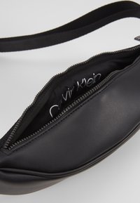 Czarna torba z sztucznej skóry o gładkiej fakturze, z zamkiem błyskawicznym i materiałowym paskiem. Wnętrze ma logo nadrukowane na czarnym wyściółce.