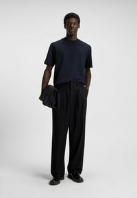 Muž stojící, oblečený v námořnickém modrém tričku, černých skládacích kalhotách, černých botách, držící v levé ruce složenou černou bundu.