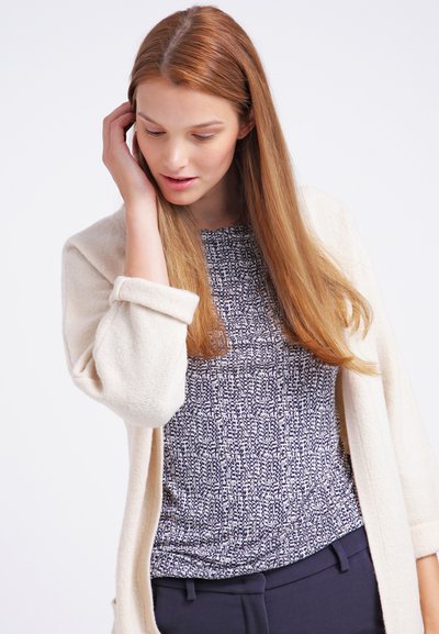 Cardigan écru sur un haut marine à motifs et à manches courtes, texture lisse, et pantalon foncé, avec des lignes simples et une coupe décontractée.