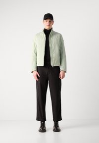 Jachetă matlasată verde deschis cu guler înalt, asortată cu un pulover negru cu guler înalt, pantaloni negri cu picioare largi și pantofi negri cu șiret. Căciulă neagră.