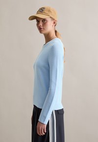 Lichtblauw shirt met lange mouwen in een gestructureerde brei, gecombineerd met een zwarte rok met witte strepen. Een beige pet maakt de look compleet.