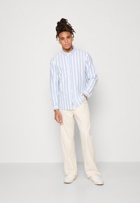 Camisa de botones a rayas azul y blanca con collar, mangas largas y pantalones anchos de color claro. El modelo está de pie frente a un fondo neutro.
