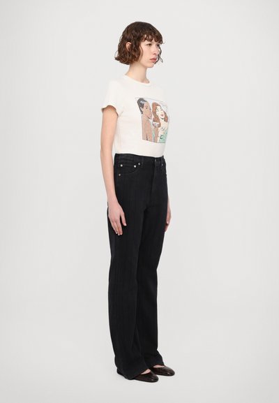 T-shirt en coton crème avec un graphique de dessin animé, associé à un jean en denim noir à rayures. Le modèle porte des ballerines noires. Fond clair.