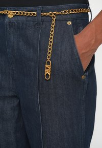 Pantalons en denim foncé avec une ceinture en chaîne dorée et un détail de fermoir. Le tissu a un tissage texturé et une coupe droite.