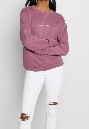 Personne portant un sweat violet avec le texte "MISSGUIDED" et un jean skinny blanc déchiré, debout, les bras croisés, devant un fond uni.