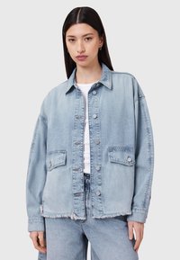 Giacca in denim chiaro con vestibilità rilassata, due tasche anteriori, bottoni a pressione in perlina e orlo sfilacciato. Abbinata a una maglietta bianca e jeans coordinati.