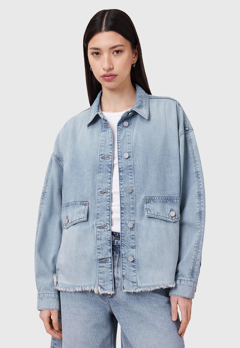 Giacca in denim chiaro con vestibilità rilassata, due tasche anteriori, bottoni a pressione in perlina e orlo sfilacciato. Abbinata a una maglietta bianca e jeans coordinati.