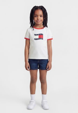 Młoda dziewczyna stoi, ubrana w białą koszulkę Tommy Hilfiger z czerwonym wykończeniem, ciemne szorty jeansowe, białe skarpetki i białe tenisówki, na jednolitym tle.