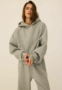 Jasnoszara, oversize bluza z kapturem i sznurkiem, żebrowane mankiety oraz miękka faktura. Zestawiona z dopasowanymi szarymi spodniami dresowymi.