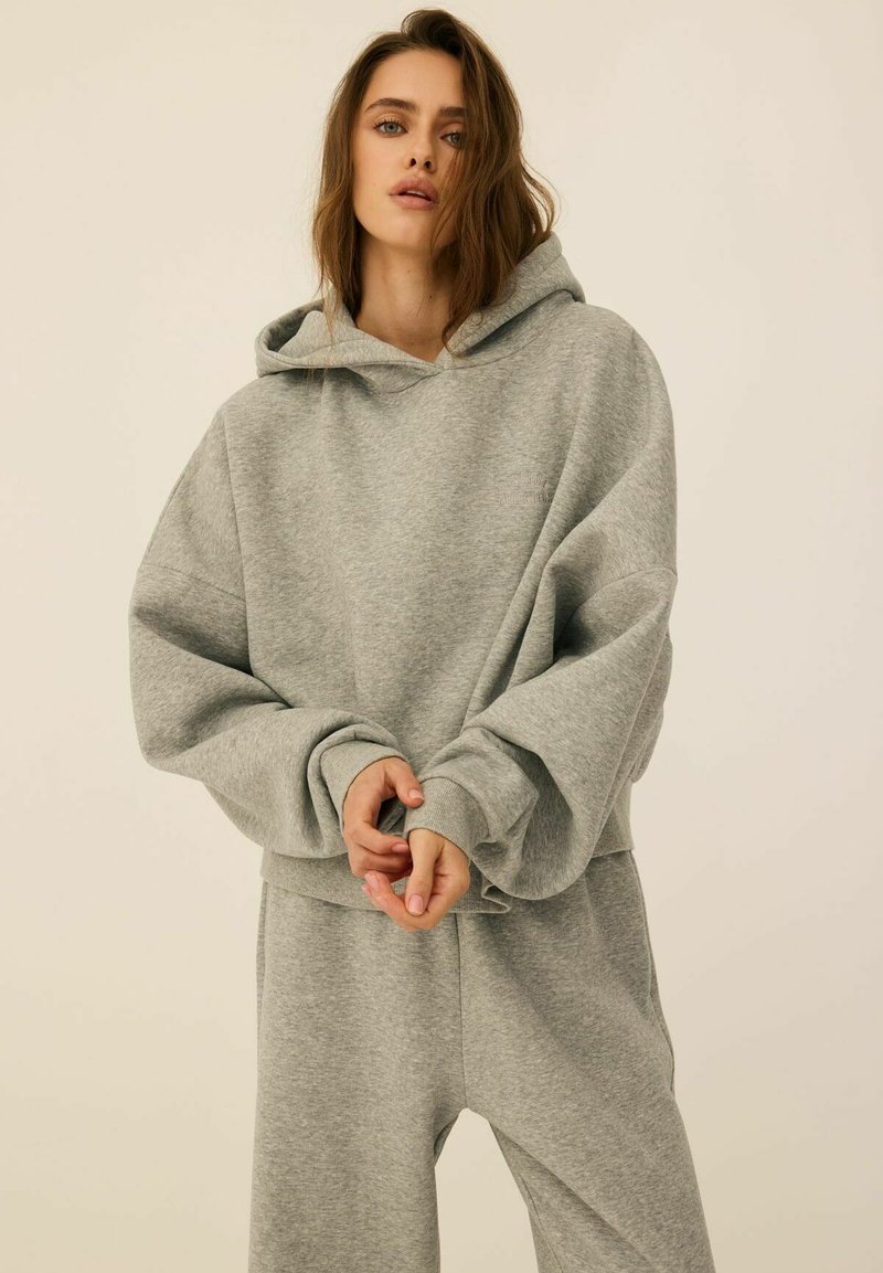 Jasnoszara, oversize bluza z kapturem i sznurkiem, żebrowane mankiety oraz miękka faktura. Zestawiona z dopasowanymi szarymi spodniami dresowymi.
