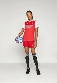 Maillot de football rouge avec des manches blanches, affichant le logo "Emirates" ; shorts rouges ; chaussettes noires avec des rayures blanches ; joueur tenant un ballon de football bleu.