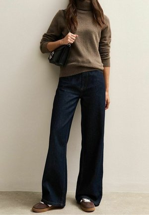 Kvinde iført brun rullekravesweater, mørke bukser med vide ben, brune sneakers og holder en sort håndtaske over skulderen.