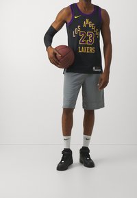 Hombre con una camiseta de baloncesto de Los Angeles Lakers sosteniendo una pelota de baloncesto, con pantalones cortos grises, zapatillas negras, calcetines blancos de Nike y una manga negra en el brazo.