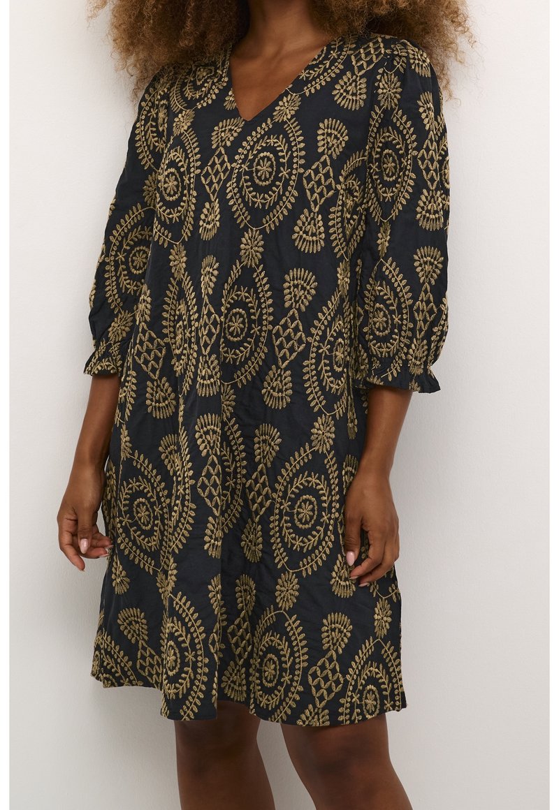 Robe noire longueur genou avec broderies dorées circulaires et florales, manches trois-quarts avec poignets élastiques, portée par une personne aux cheveux bouclés.