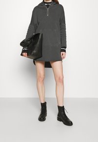 Person som bär en mörkgrå oversized hoodieklänning, svarta snörade ankelstövlar, och håller en stor svart vävd läderhandväska.