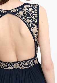 Marinblå klänning med öppen rygg, prydd med intrikata gyllene blommönster längs axlarna och midjan, samt en plisserad kjoldetekstur.