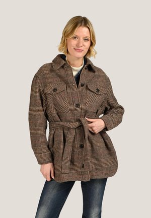 Femme en veste marron à carreaux ceinturée avec poches poitrine boutonnées, portant un jean foncé, debout devant un fond uni.