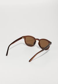 Braun getönte Sonnenbrille mit runden Gläsern, leichtgewichtigen transparenten Rahmen und schlanken Bügeln mit dezentem Branding.