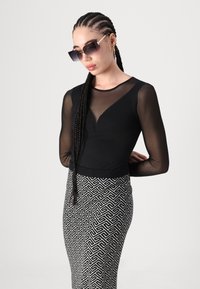 Haut noir avec des manches transparentes et un col en V ; jupe noir et blanc à motifs avec un design géométrique ; lunettes de soleil oversize ; cheveux longs tressés.