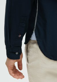 Knopfhemd aus marineblauem Stoff mit strukturiertem Gewebe; verfügt über kontrastierende weiße Nähte und markenprägte Knöpfe; seitlicher Schlitz zeigt gestreiften Stoff.