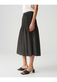 Jupe midi noire avec un corsage smocké à motifs et un ourlet évasé, associée à des mules en cuir noir à bout ouvert sur un fond clair.