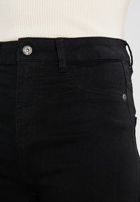 Vaqueros de mezclilla negra de cintura alta con cremallera, botón plateado, bolsillos frontales y detalles de costura sutiles. Textura suave y diseño ajustado.