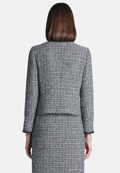 Femme aux cheveux bruns raides portant une veste texturée noire et blanche à manches longues et une jupe assortie, debout de dos.
