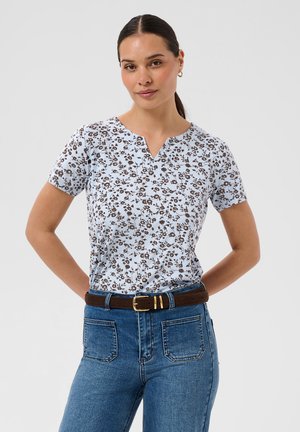 Femme aux cheveux foncés attachés en queue de cheval, portant un haut bleu clair à fleurs à manches courtes, un jean bleu et une ceinture marron, debout les mains sur les hanches.