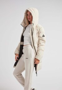 Chaqueta de plumas color crema con una gran capucha, cierre frontal y bolsillos laterales, combinada con joggers de un ligero color beige que presentan un pequeño logo.