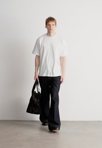 Λευκό κοντομάνικο μπλουζάκι με διακριτό λογότυπο στο στήθος, σε συνδυασμό με σκούρες φαρδιές τζιν παντελόνες και κρατώντας μια μαύρη τσάντα ώμου.