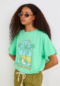T-shirt vert menthe à manches courtes avec un graphique tropical représentant des palmiers et une plage, associé à un short à cordon de serrage vert olive.