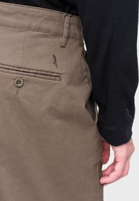 Vaaleat puuvillaiset housut, joissa on takatasku, nappikiinnitys ja ommellut yksityiskohdat. Rintataskun ylle on kirjailtu hillitty logo.