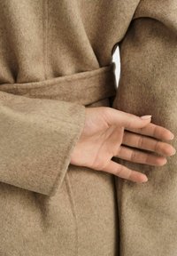 Beżowy wełniany szlafrok z przewiązanym paskiem, eksponujący miękką teksturę i zbliżenie dłoni sięgającej za tkaninę.