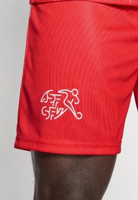 Piros atlétikai rövidnadrág texturált felülettel, fehér grafikus logóval és felirattal az oldalon, rugalmas derékrésszel, zsebek nélkül.